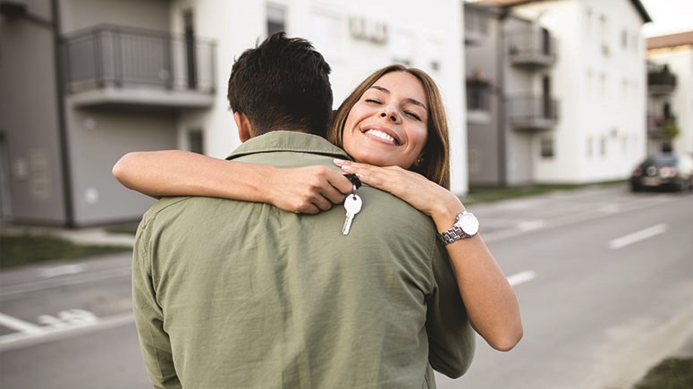 Young couple hugging, she's holding a house key, they're on a street in front of an apartment building